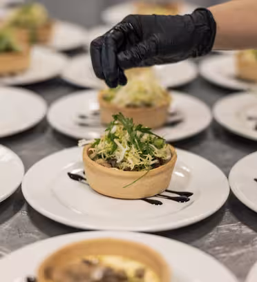 A chef wearing a black glove garnishes a tart with greens on a white plate. The tart is filled with a savory mix and drizzled with sauce. Other identical dishes are arranged on the table in the background.