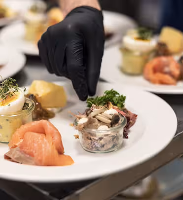 A chef wearing black gloves is garnishing plates of gourmet food. The dishes include smoked salmon, cheese, pickles, a small jar of salad with mushrooms, and boiled eggs. Plates are arranged neatly on a metal surface.