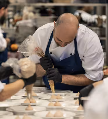 A chef wearing a blue apron pipes cream onto plates in a busy kitchen, surrounded by fellow chefs in white uniforms focused on preparing dishes. Culinary work is in progress with trays and ingredients visible in the background.