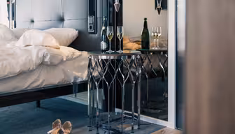 A modern bedroom with an unmade bed, a pair of sandals on the floor, and a small round table adorned with two glasses of white wine and a wine bottle. A mirror on the right reflects the table and drinks.