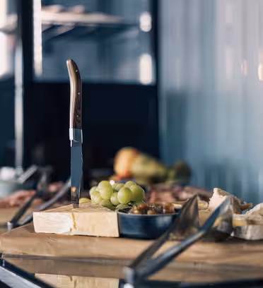 A kitchen counter with a variety of cheeses, a knife embedded in one block, surrounded by green grapes. A metal dish and tongs are nearby. Blurred background with shelves and kitchen items.