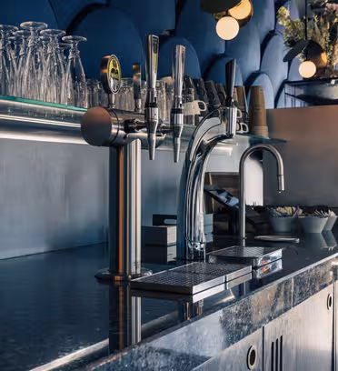 Bar counter with a glossy black surface, featuring beer taps, an espresso machine, and shelves with glasses. The backdrop includes a blue, textured wall and soft lighting.