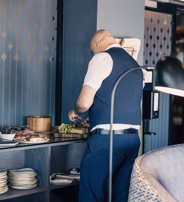 A man in a blue vest stands at a buffet table, arranging food such as grapes and cold cuts. Plates and serving dishes are neatly placed nearby. The setting appears to be a restaurant or hotel dining area with modern decor.