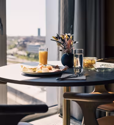 A small dining table set for one person with a plate of breakfast foods, a glass of orange juice, and water. A vase with flowers decorates the table. The setting is by a window overlooking a cityscape under clear skies.