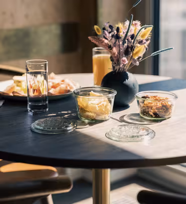 A round table with a small vase of dried flowers, two glasses filled with water and orange juice, covered glass bowls with snacks, and a plate with sliced bread. Sunlight streams through a nearby window, creating a cozy ambiance.