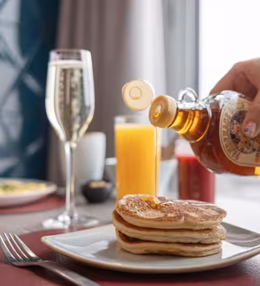 A hand pours syrup over a stack of pancakes on a white plate. Nearby, theres a glass of champagne, a glass of orange juice, and a cup of coffee on a wooden table. A fork and knife are placed on a red placemat.