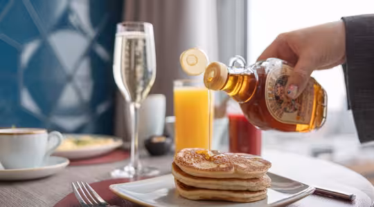A hand pours syrup over a stack of pancakes on a white plate. Nearby, theres a glass of champagne, a glass of orange juice, and a cup of coffee on a wooden table. A fork and knife are placed on a red placemat.