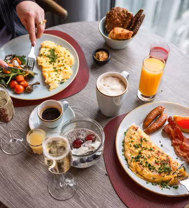 Two people are having a meal at a round table by a window. Plates with omelets, sausages, bacon, and salad are accompanied by drinks, including coffee, orange juice, and champagne. A bowl of yogurt with fruit and pastries are also on the table.