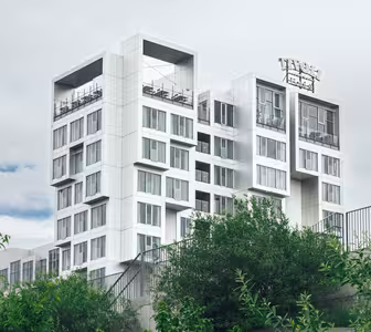 Modern multilevel white building with square design elements, featuring glass walls and rooftops. Surrounded by lush greenery, it stands against a cloudy sky. Signage is partially visible on the top level.