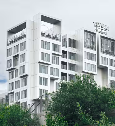 Modern multilevel white building with square design elements, featuring glass walls and rooftops. Surrounded by lush greenery, it stands against a cloudy sky. Signage is partially visible on the top level.