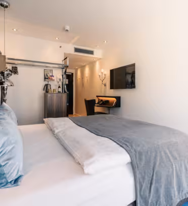 Modern hotel room with a large bed featuring white linens and a gray blanket. A flat-screen TV is mounted on the wall. There is a small desk with a chair, a kitchenette area with a minibar, and soft lighting creating a cozy atmosphere.