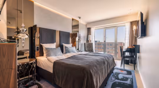 A modern hotel room featuring a large double bed with gray bedding and pillows. The room has mirrored walls, a desk with a chair, a mounted flat-screen TV, and large windows with city views. Elegant lighting fixtures hang on either side of the bed.