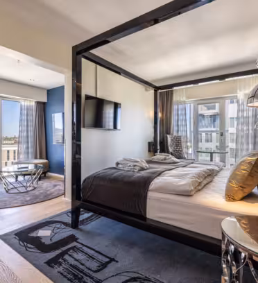 Luxurious modern hotel room featuring a canopy bed with black frame and white bedding. Large windows reveal a city view. A sitting area with a round glass table and chairs is in the corner. Decorative silver lamps and complimentary water bottles are present.