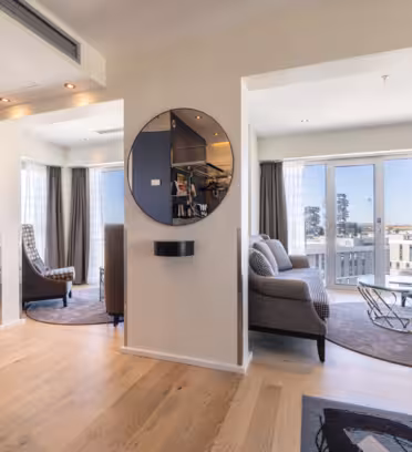 Modern living room with light wood flooring, neutral-colored sofas, and a glass coffee table. Large windows let in natural light. A round mirror and decorative lighting add elegance to the room. A kitchenette is visible in the foreground.