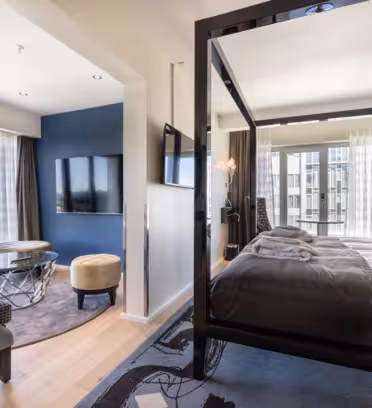 A modern hotel room with a four-poster bed covered in dark gray and white bedding. A wall-mounted TV and two round chairs surround a small glass table in a sitting area. Large windows provide ample natural light and city views.
