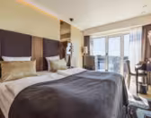 Modern hotel room with a large bed featuring brown and gold bedding, a stylish headboard, and bedside lamps. A floor-to-ceiling window offers a city view, and a desk with a chair sits to the right, creating a bright, inviting atmosphere.
