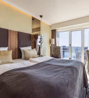 Modern hotel room with a large bed featuring brown and gold bedding, a stylish headboard, and bedside lamps. A floor-to-ceiling window offers a city view, and a desk with a chair sits to the right, creating a bright, inviting atmosphere.