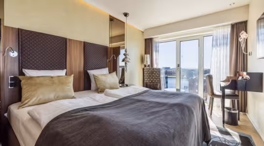 Modern hotel room with a large bed featuring brown and gold bedding, a stylish headboard, and bedside lamps. A floor-to-ceiling window offers a city view, and a desk with a chair sits to the right, creating a bright, inviting atmosphere.