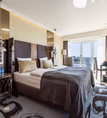 Modern hotel room with a large bed, dark headboard, and stylish bedside lamps. Floor-to-ceiling windows offer a city view. A desk with a chair is on the right. Neutral tones and contemporary decor create a sleek, inviting atmosphere.