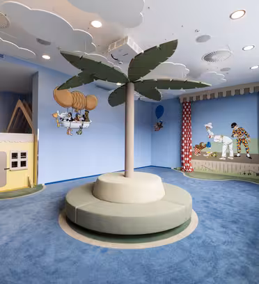 A colorful childrens playroom with a large, central palm tree sculpture surrounded by seating. The walls have playful murals featuring characters and a small playhouse with red accents. The ceiling is decorated with hanging cloud shapes.