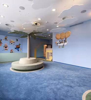 A childrens playroom with light blue walls, featuring cartoon characters on the walls. The ceiling is decorated with clouds, and a central tree-shaped structure adds a playful touch. The room has soft, cushioned flooring.