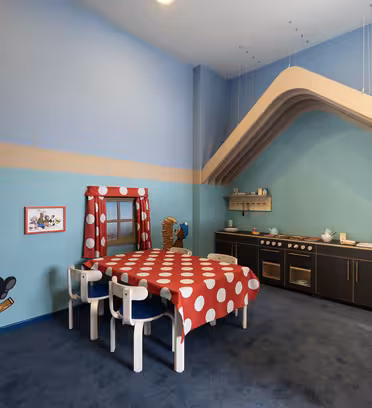 A playful room with a childrens kitchen set against a blue wall. The dining table is covered with a red and white polka dot cloth, surrounded by small chairs. Colorful murals of cartoon animals decorate the walls. The carpet is deep blue.