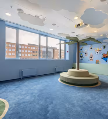 A childrens playroom with a nautical theme featuring a lighthouse, blue carpet resembling water, and a round seating area with a palm tree. Walls are painted with clouds, butterflies, and cartoon characters. Large windows let in natural light.