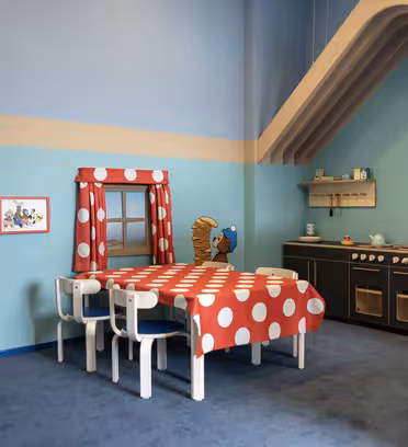 A playful kitchen and dining area with a red polka dot tablecloth, small white chairs, a cartoon bear on the wall, and child-sized wooden kitchen units. The room features bright blue and teal walls with whimsical decor.