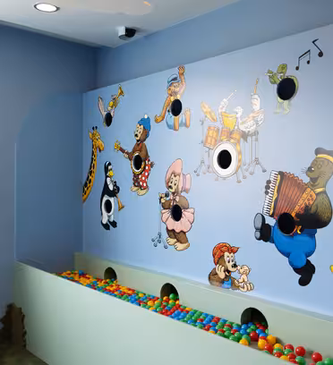 A colorful childrens playroom wall featuring cartoon animal characters playing musical instruments. Below are holes and a pit filled with multicolored balls. The walls and environment have a cheerful, playful design.
