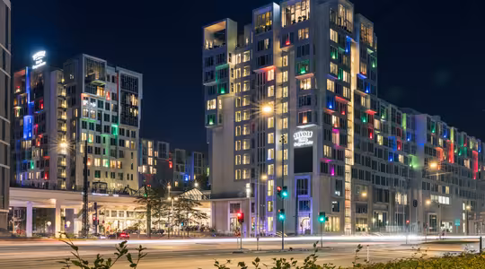 Night view of a modern hotel building with illuminated windows in various colors, creating a vibrant pattern. Streetlights and car trails are visible on the road in front, adding dynamic motion to the scene.