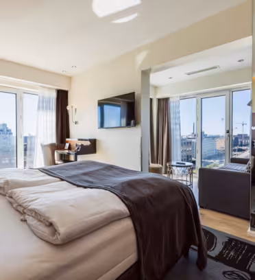 Modern hotel room with a large bed, dark bedding, and patterned cushions. A TV is mounted on the wall opposite the bed. Floor-to-ceiling windows provide a city view. Theres a small seating area with chairs and a table near the window.