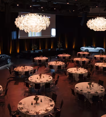 A dimly lit banquet hall with round tables set for an event. Two cars are displayed on either side, and a large screen is on stage. Elegant chandeliers hang from the ceiling, and warm orange lights illuminate the walls.