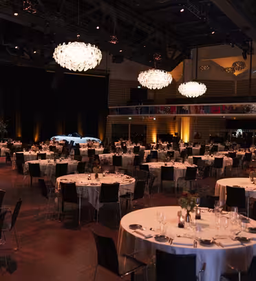 A spacious event hall with round tables set for a formal dinner, featuring white tablecloths and elegant place settings. Large chandeliers hang from the ceiling, and a screen with the text Van Mossel is visible. Warm lighting creates an inviting atmosphere.