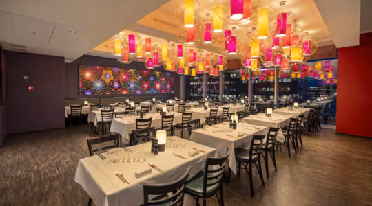 A spacious, modern restaurant with neatly arranged tables covered in white cloths. Colorful lanterns hang from the ceiling, and large windows reveal a cityscape. A vibrant flower-patterned wall adds to the lively ambiance.