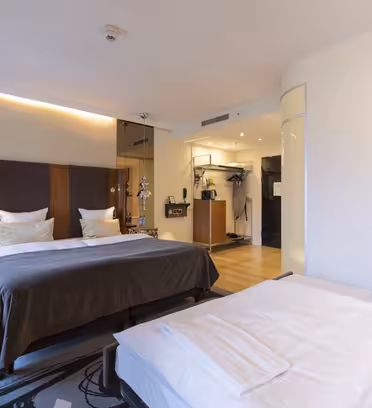 Modern hotel room featuring two beds with white linens and dark blankets, soft lighting, and neutral decor. A wall with artistic designs, pendant lights, a flat-screen TV, and a workstation are visible. The floor is wooden, and theres an abstract-patterned rug.