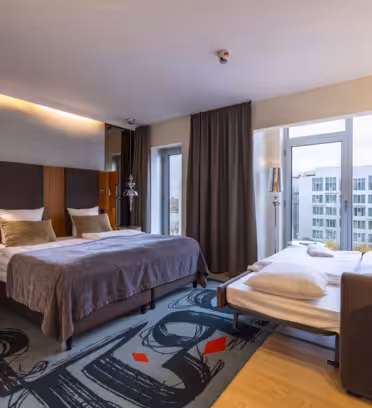 Hotel room with two beds: a double and a single. The room features a modern design, with a patterned carpet, a large window with city views, dark curtains, and contemporary lighting. A wooden desk and chair are near the window.