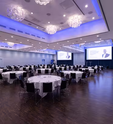 A large conference room with round tables and chairs arranged neatly, set for an event. Two large screens display a presentation slide, and the ceiling features modern lighting fixtures with blue accents along the edges.