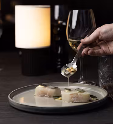 A hand drizzles sauce over two pieces of fish topped with small cucumber slices on a plate. The table is set with wine glasses, a water glass, and a lit cylindrical lamp, creating a cozy dining ambiance.