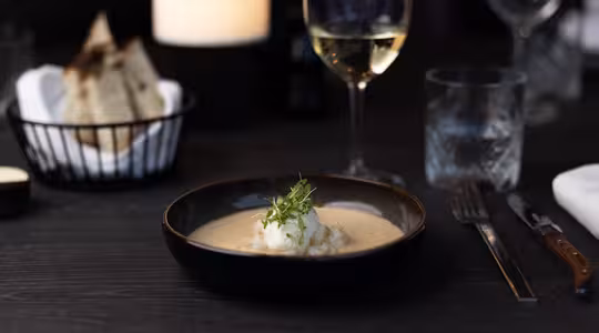A stylish dinner with a bowl of soup garnished with vegetables in the foreground. A glass of white wine stands next to it. In the background you can see a basket of sliced bread and a gently burning candle.