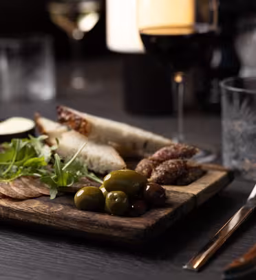 A wooden board with assorted appetizers, including sliced bread, cured meats, cheeses, olives, and arugula, is on a dark table. A glass of red wine and another glass are alongside, with utensils and a napkin nearby.