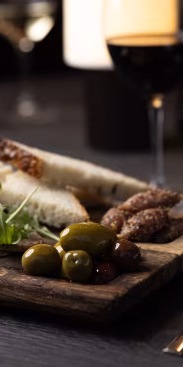 A wooden board with assorted appetizers, including sliced bread, cured meats, cheeses, olives, and arugula, is on a dark table. A glass of red wine and another glass are alongside, with utensils and a napkin nearby.