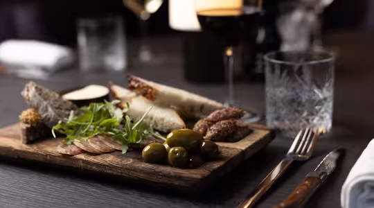 A wooden board with assorted appetizers, including sliced bread, cured meats, cheeses, olives, and arugula, is on a dark table. A glass of red wine and another glass are alongside, with utensils and a napkin nearby.