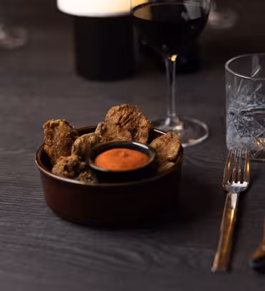 A bowl of fried mushrooms with a small dish of dipping sauce sits on a dark wooden table. Nearby are a glass of red wine, an empty textured glass, and a knife and fork. The background includes another glass and a dim candle.
