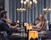 A family of four sits at a dimly lit restaurant table. The parents face each other, while two young boys sit between them. Elegant chandeliers hang above, and soft lighting creates a cozy atmosphere.