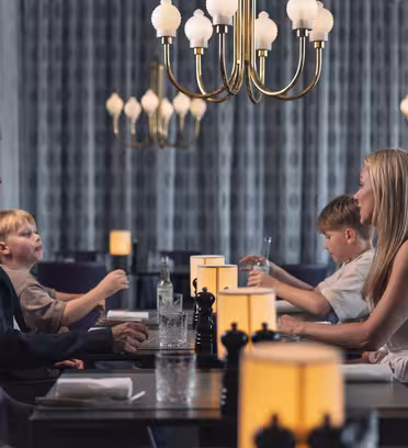 A family of four sits at a dimly lit restaurant table. The parents face each other, while two young boys sit between them. Elegant chandeliers hang above, and soft lighting creates a cozy atmosphere.