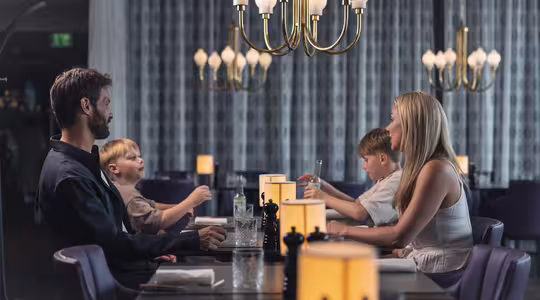 A family of four sits at a dimly lit restaurant table. The parents face each other, while two young boys sit between them. Elegant chandeliers hang above, and soft lighting creates a cozy atmosphere.