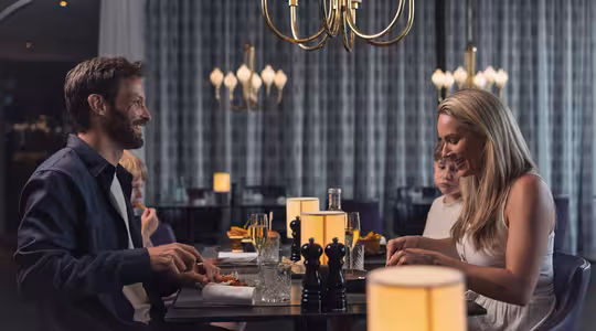 A couple sits across from each other at a restaurant table, smiling and engaging in conversation. The man is on the left, the woman on the right. The room is softly lit, with elegant chandeliers and a patterned curtain in the background.