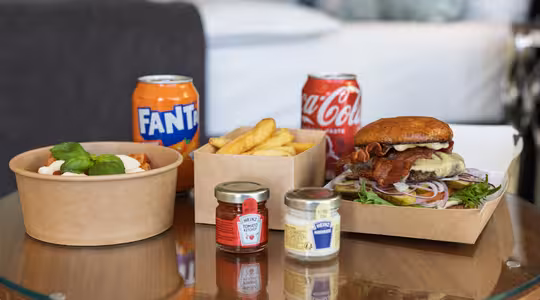 A hotel room service meal on a glass table includes a cheeseburger with bacon and lettuce in a takeaway box, fries, pasta in a bowl with basil garnish, two small condiment jars, and cans of Fanta and Coca-Cola against a cozy room background.