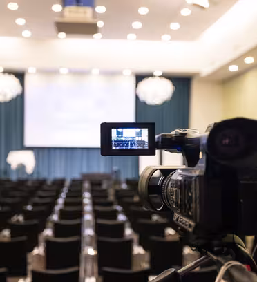 A video camera on a tripod records an empty conference room with rows of black chairs facing a blue stage with a podium. Bright overhead lights illuminate the room, and a blurry projection screen is visible in the background.