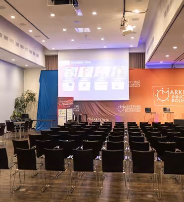 An empty meeting room set up for a marketing event. Rows of black chairs face a stage with a podium and large screens displaying speaker information. Banners read Marketing Innovation Roundtable. Video equipment is set up in the back.